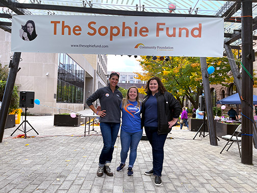 Visions AVP/Chief Governance and Strategy Officer, Jenna stands for a picture at the Ithaca Commons under a banner reading Sophie's Fund with Community Development Liaisons, Sarah and Jocelyn.