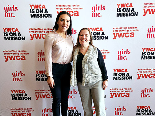 Visions employees Kendra (left) and Sarah (right) pictured in front of a wall containing the following text: “Girls Inc,” “YWCA is on a Mission,” and “Eliminating Racism/Empowering Women YWCA.”